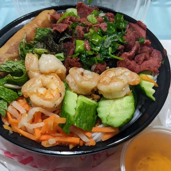 A Vietnamese vermicelli bowl with shrimp, beef, egg roll, and vegetables in a black bowl.