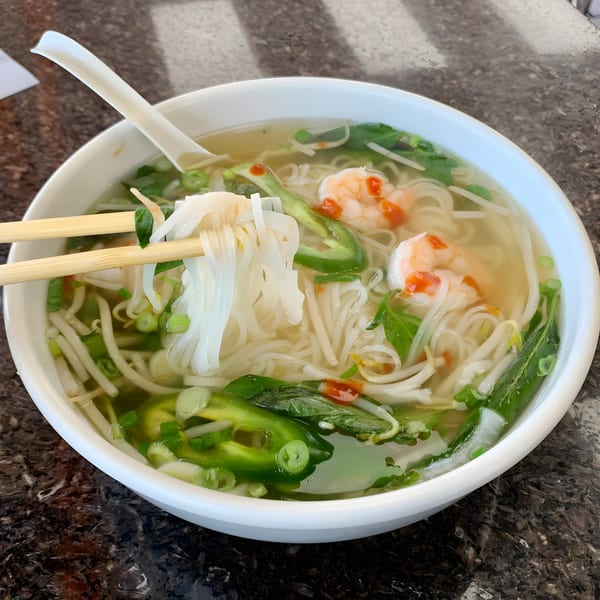 Chopsticks lift white rice noodles from a bowl of BMC Pho with shrimp, jalapeños, and herbs.