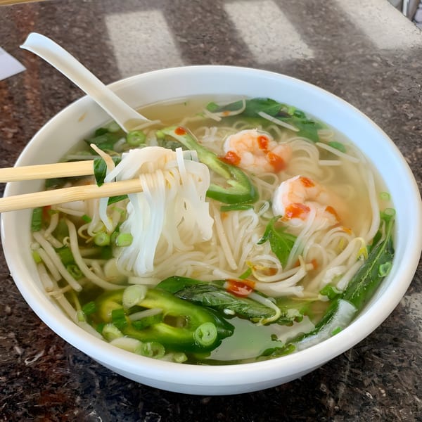 Chopsticks lifting noodles from a bowl of BMC Pho with shrimp, jalapeños, and bean sprouts.