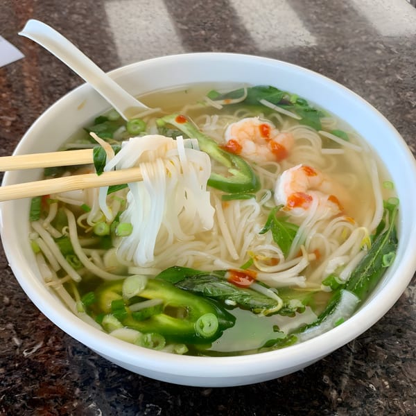 Chopsticks lifting pho noodles from a bowl with shrimp, jalapeños, and bean sprouts.