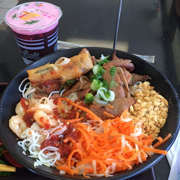 A Vietnamese vermicelli bowl with grilled pork, shrimp, egg roll, peanuts, and shredded carrots, with a pink drink.
