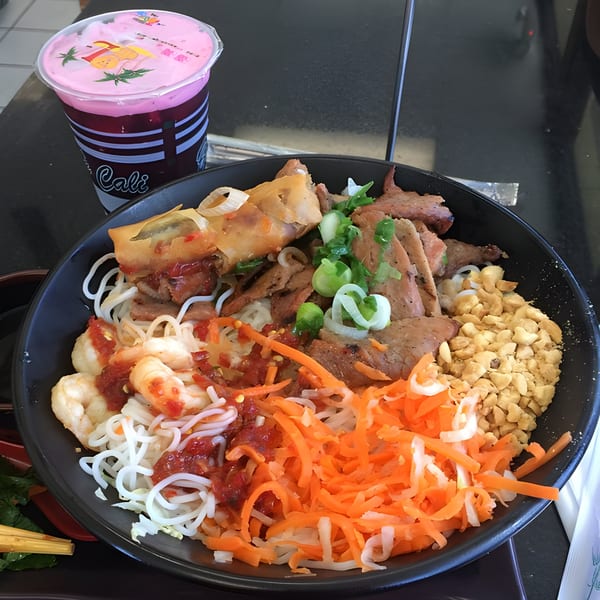 A large bowl of Vietnamese vermicelli noodles with grilled pork, shrimp, egg rolls, and vegetables, with a pink drink.