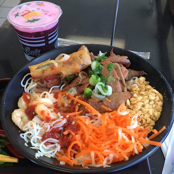 Vietnamese vermicelli bowl with grilled pork, shrimp, egg roll, peanuts, and shredded carrots, with a pink drink.