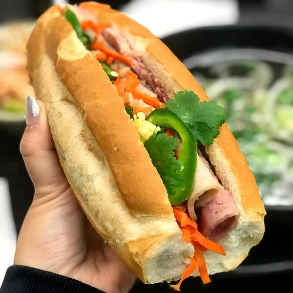 A hand holds a Vietnamese banh mi sandwich filled with meat, pickled carrots, jalapeños, and cilantro.