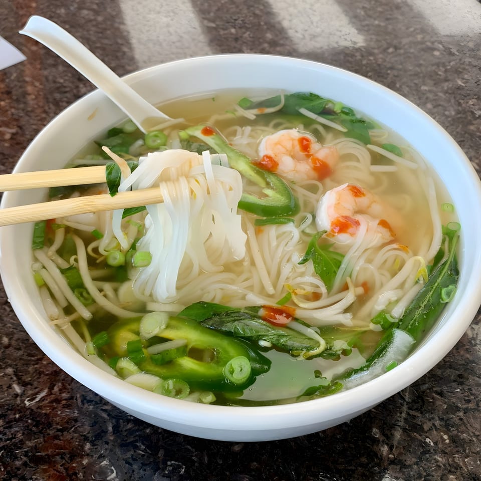 Chopsticks lifting noodles from a bowl of BMC Pho with shrimp, jalapeños, and herbs.