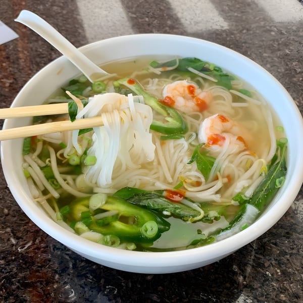 Chopsticks lifting pho noodles from a bowl with shrimp, jalapeños, and bean sprouts.