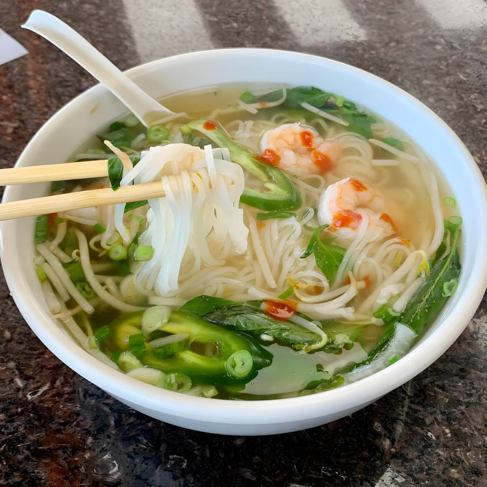 Chopsticks lifting noodles from a bowl of BMC Pho with shrimp, jalapeños, and bean sprouts.