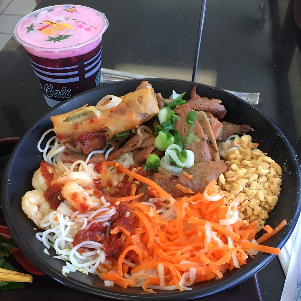 A Vietnamese vermicelli bowl with shrimp, pork, egg roll, peanuts, and shredded carrots, with a pink drink.
